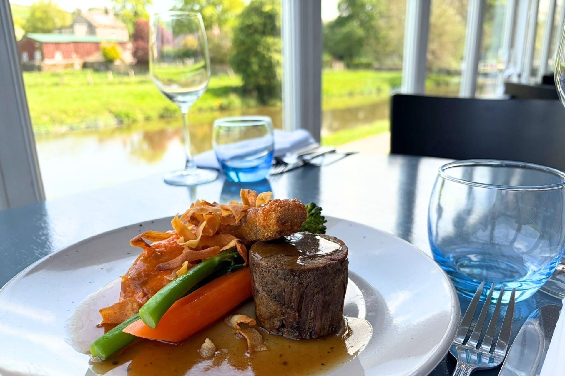 A plate of food with a steak, vegetables, and sauce, a glass of water, and a wine glass on a table with a view of greenery and buildings.