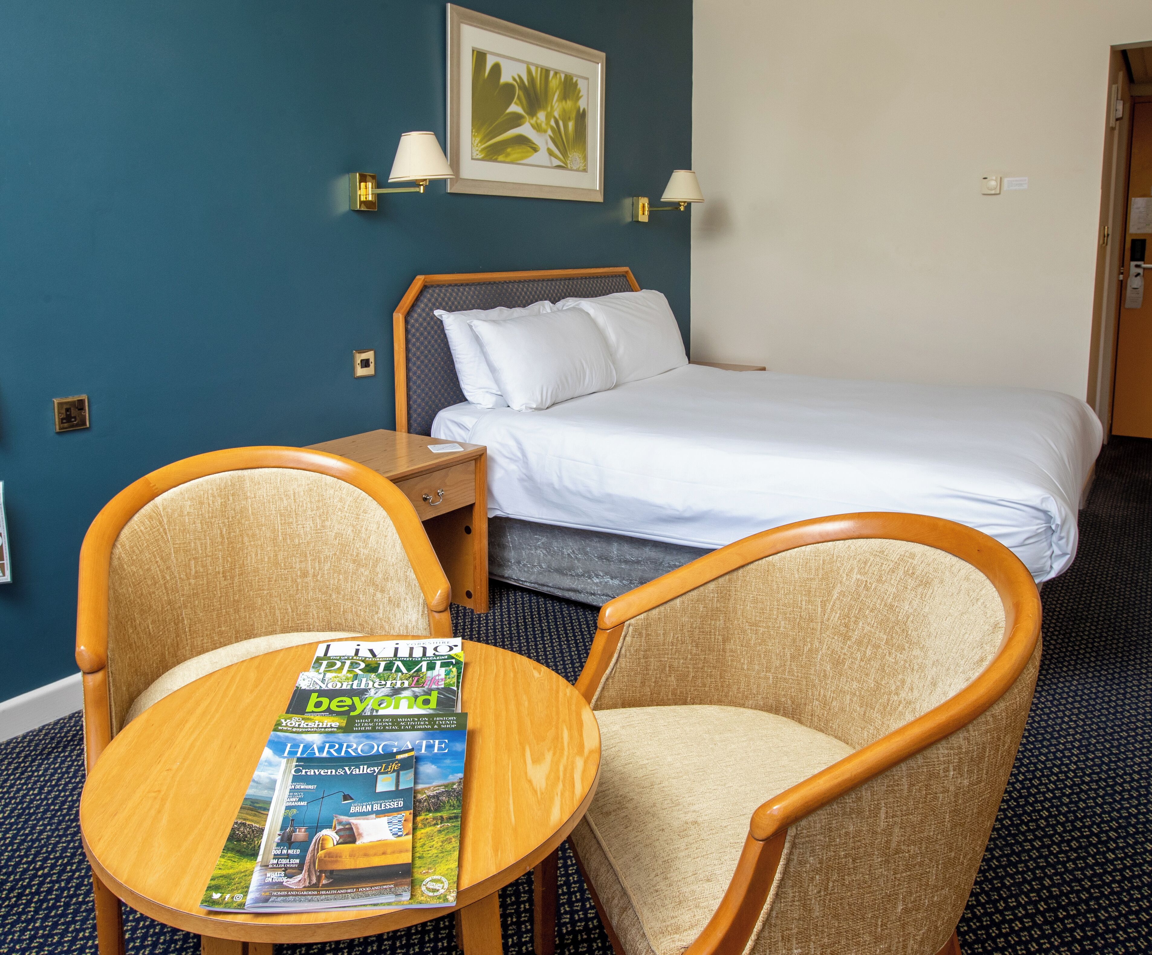 Une chambre d’hôtel avec un lit, deux fauteuils, une table en bois avec des magazines et un tableau encadré au mur.