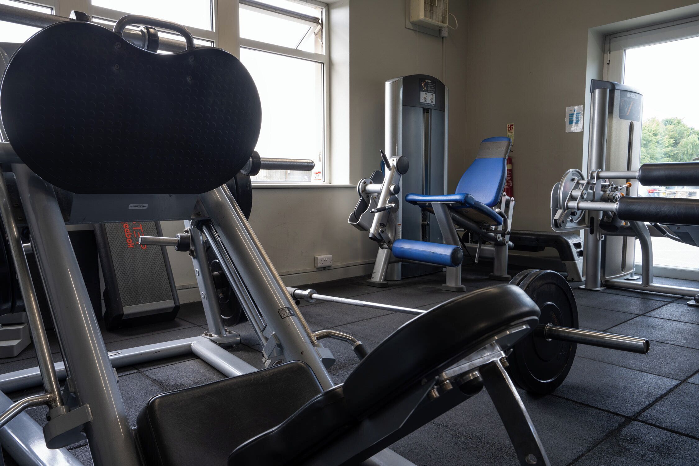 Een sportschool met diverse fitnessapparaten, waaronder een bankdrukpers, een beendrukapparaat en een leg press voor het zitten.