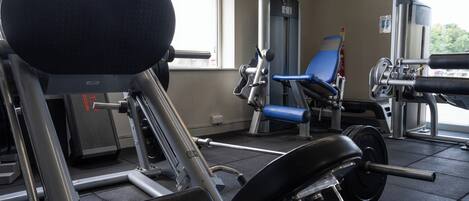 Een sportschool met diverse fitnessapparaten, waaronder een bankdrukpers, een beendrukapparaat en een leg press voor het zitten.