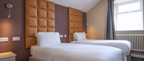 A hotel room with two beds, a brown padded headboard, a window with curtains, and a bedside table with a lamp.