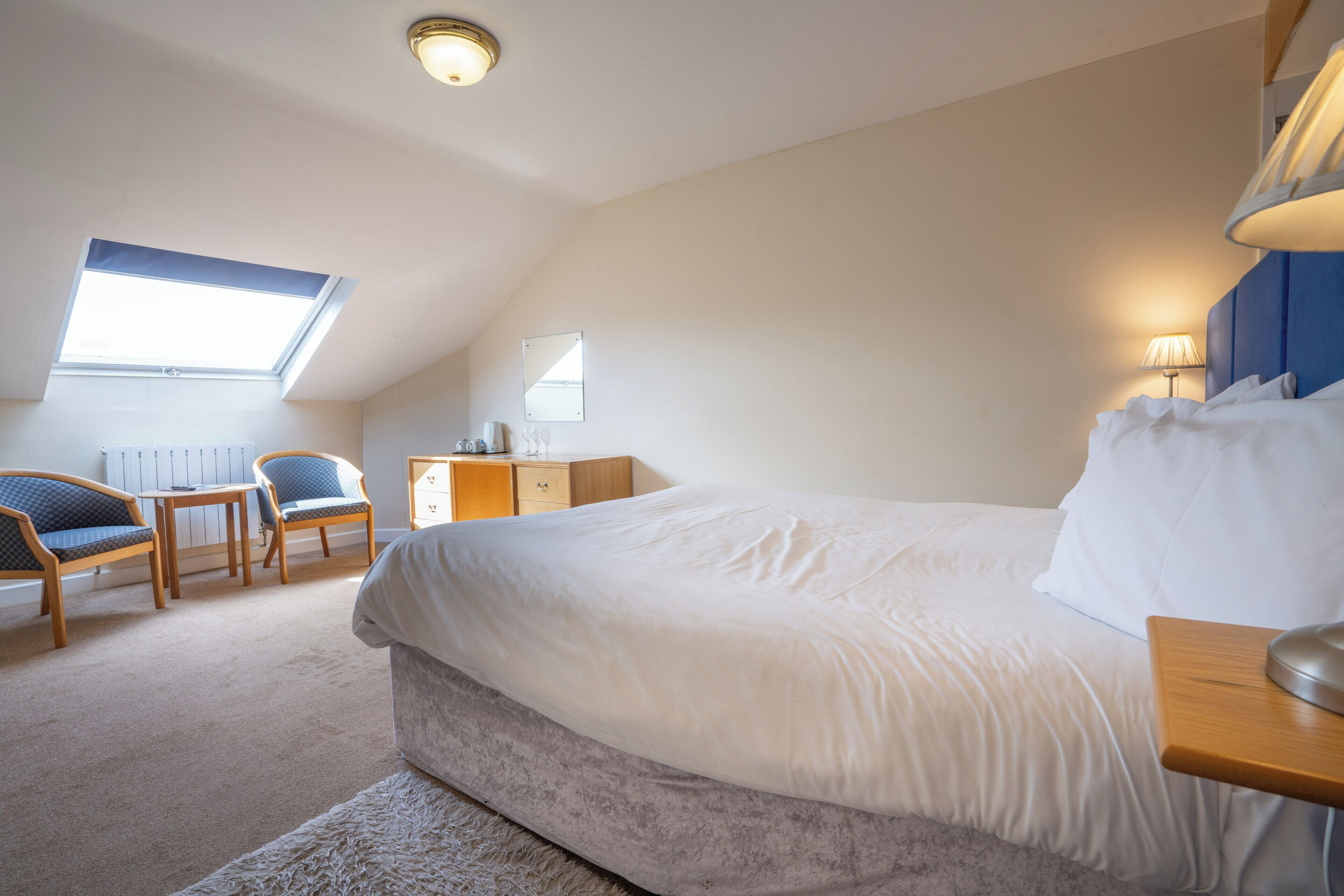 A bedroom with a large bed, a desk, two chairs, a window, and a lamp.