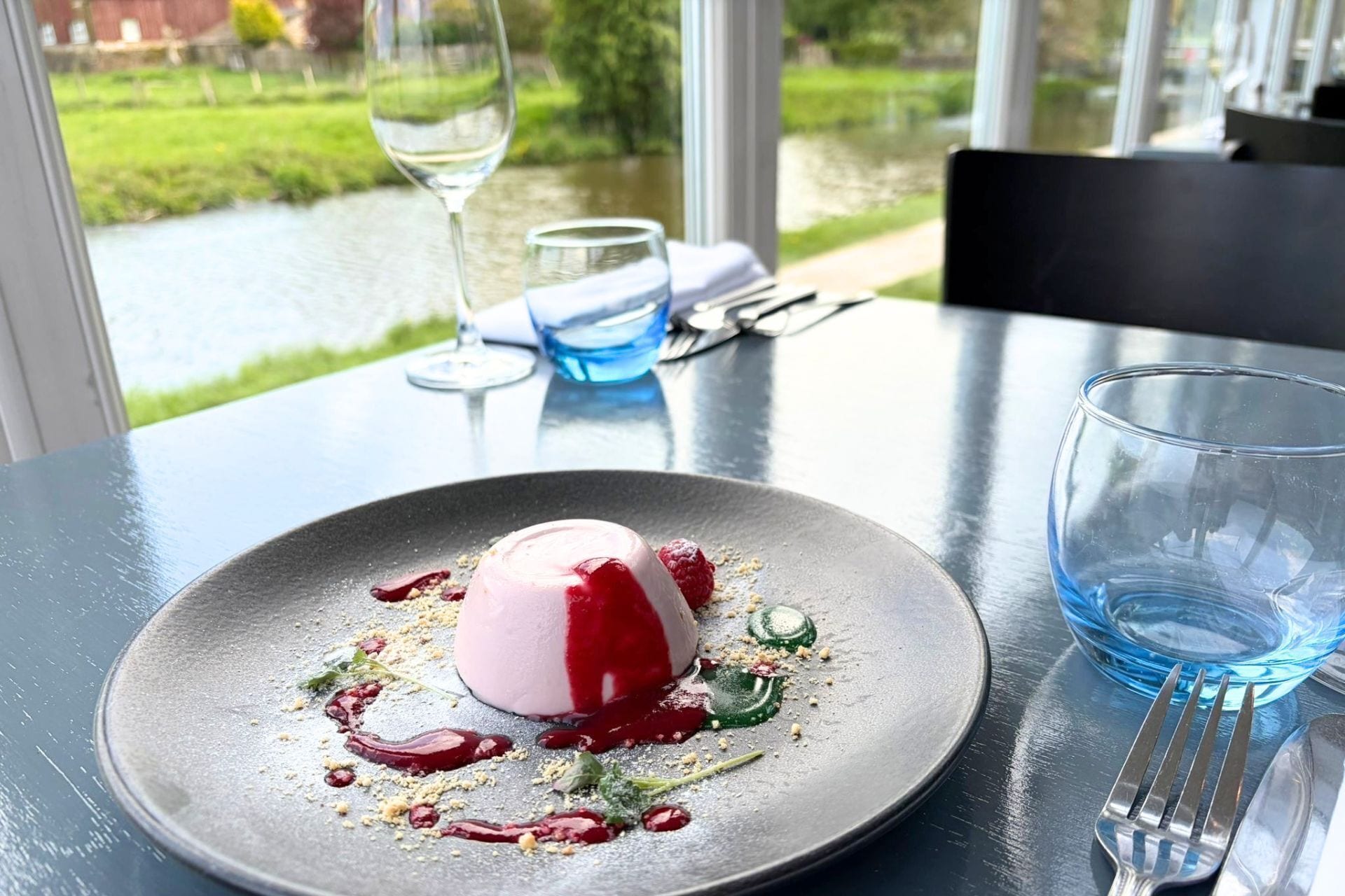 A dessert on a plate with a glass of water and a fork on a table with a view of a river.