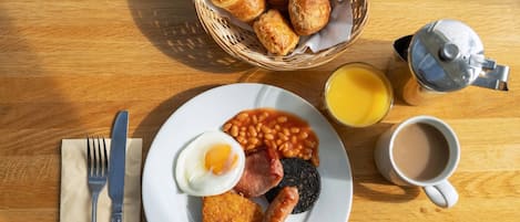 A breakfast plate with eggs, beans, bacon, sausages, and toast, accompanied by a coffee cup and a glass of orange juice on a wooden table.
