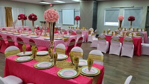 Ein Festsaal mit gedeckten Tischen für eine festliche Veranstaltung, mit rosa und goldenen Tischdecken, weißen Stühlen mit goldenen Bändern und floralen Mittelpunkten.