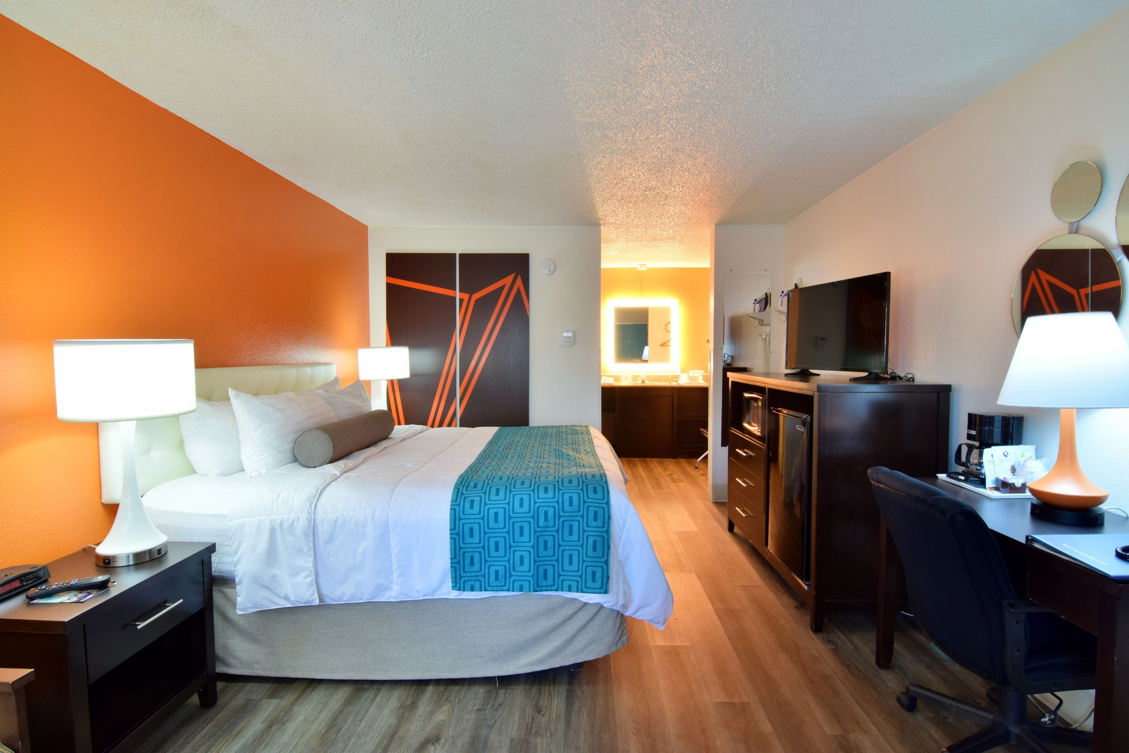 A hotel room with a bed, bedside tables, a desk, a chair, a TV, a mirror, and a bathroom with a shower and toilet.