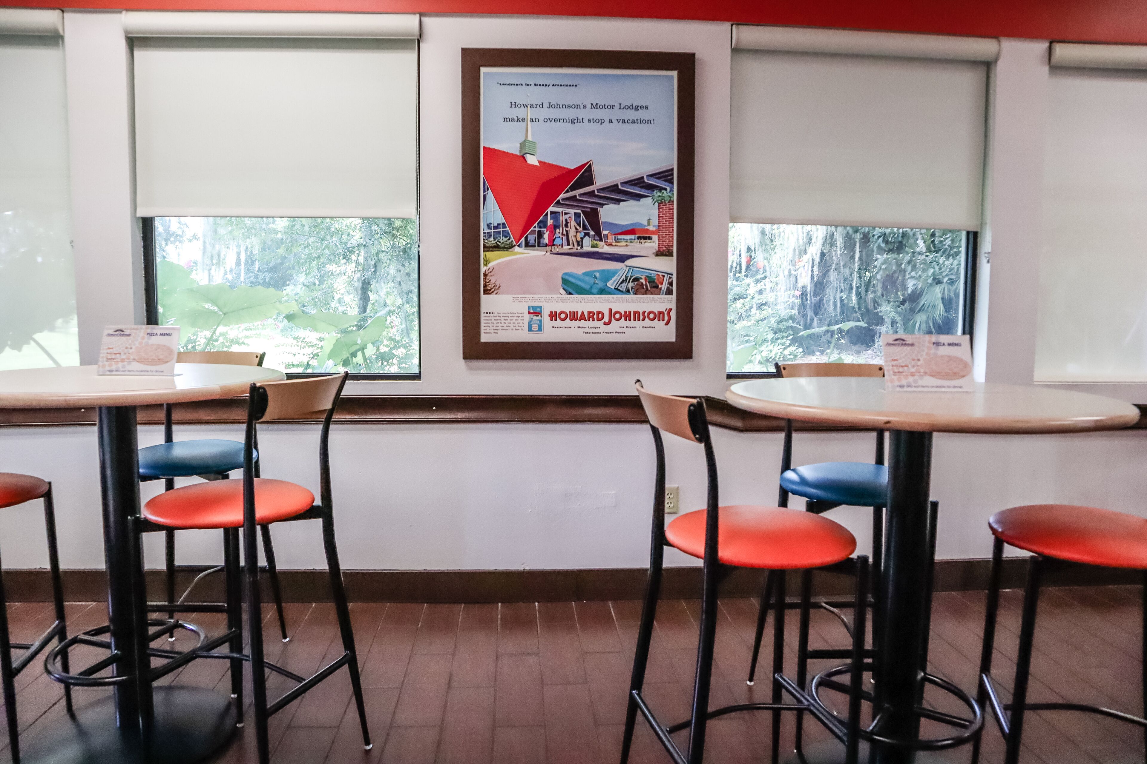 A restaurant interior with two tables and four chairs, a framed advertisement on the wall, and windows with a view of greenery outside.