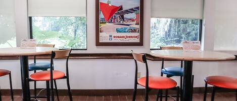 A restaurant interior with two tables and four chairs, a framed advertisement on the wall, and windows with a view of greenery outside.