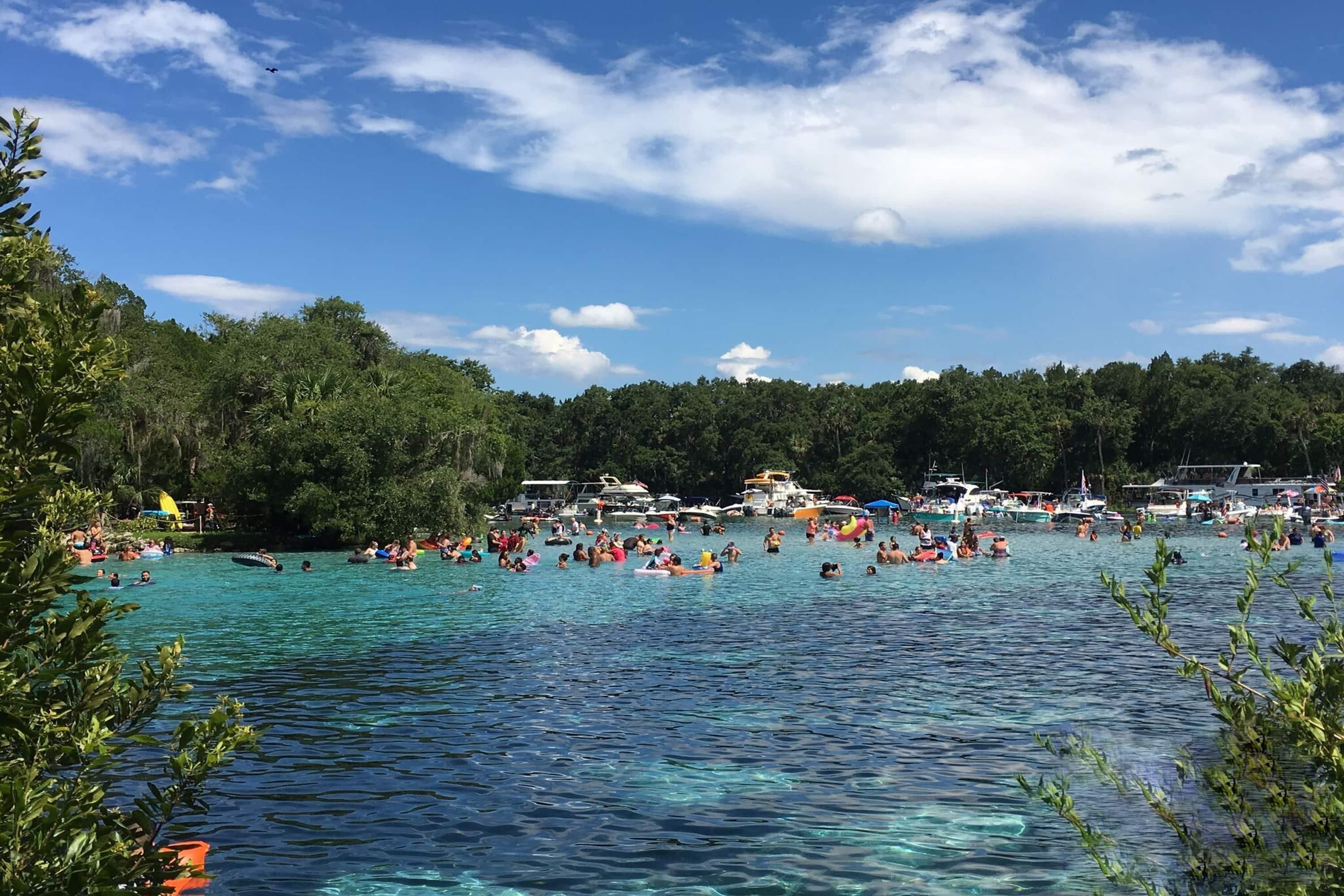 A crowded beach with clear water and boats.