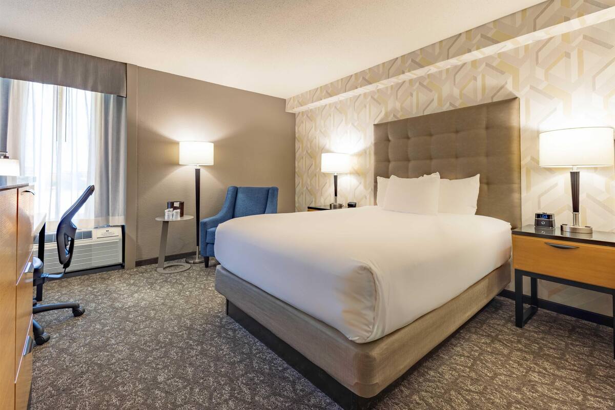 A hotel room with a large bed, two bedside tables with lamps, a desk, a chair, and a window with curtains.