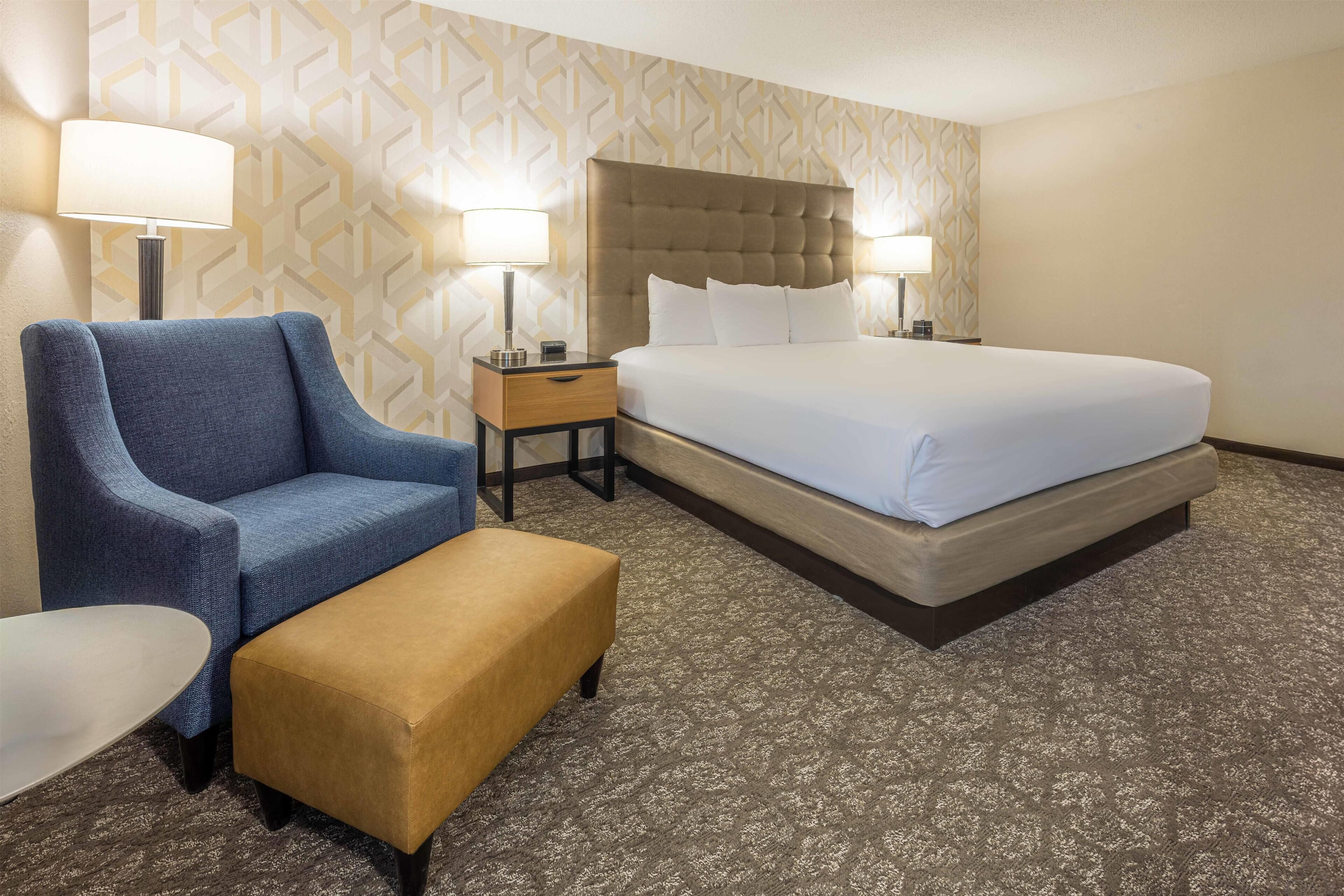 Una habitación de hotel con una cama, un sillón azul, una mesita, un banco y dos lámparas de noche.