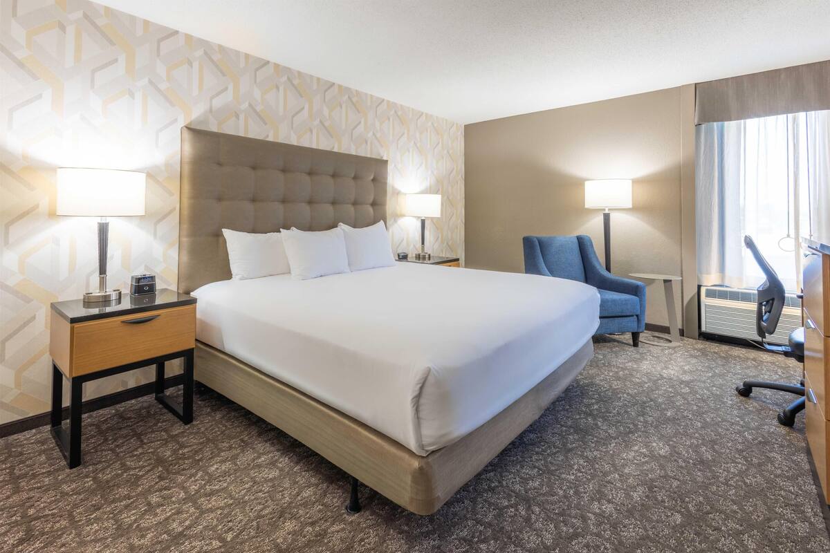 A hotel room with a large bed, two bedside tables with lamps, a blue armchair, and a view of the outdoors.