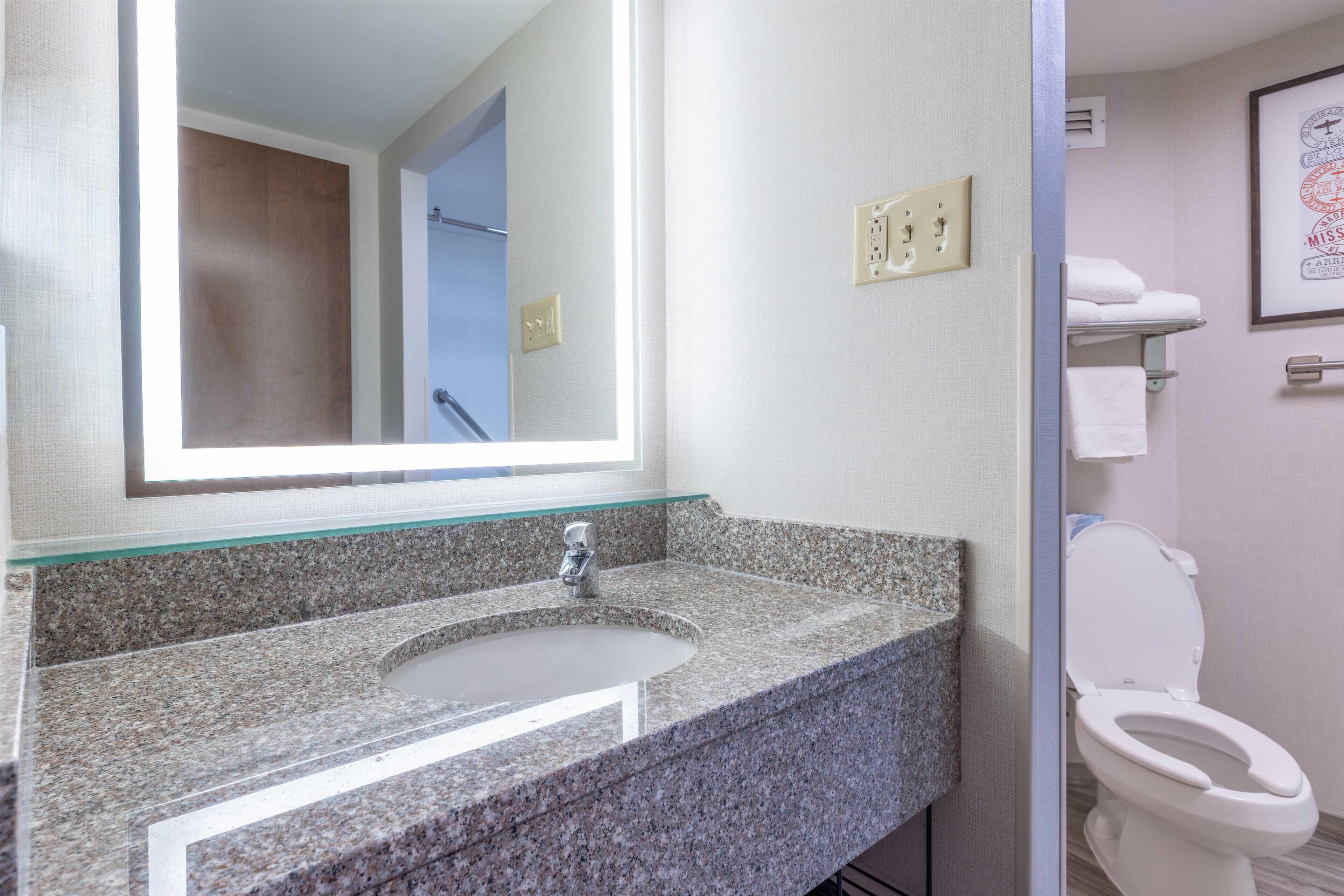 A bathroom with a granite countertop sink, a mirror, a white toilet, and a framed picture on the wall.