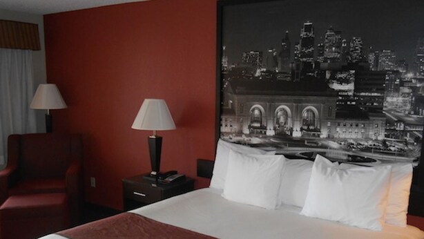 A hotel room with a large bed, a red wall, and a cityscape painting above the bed.