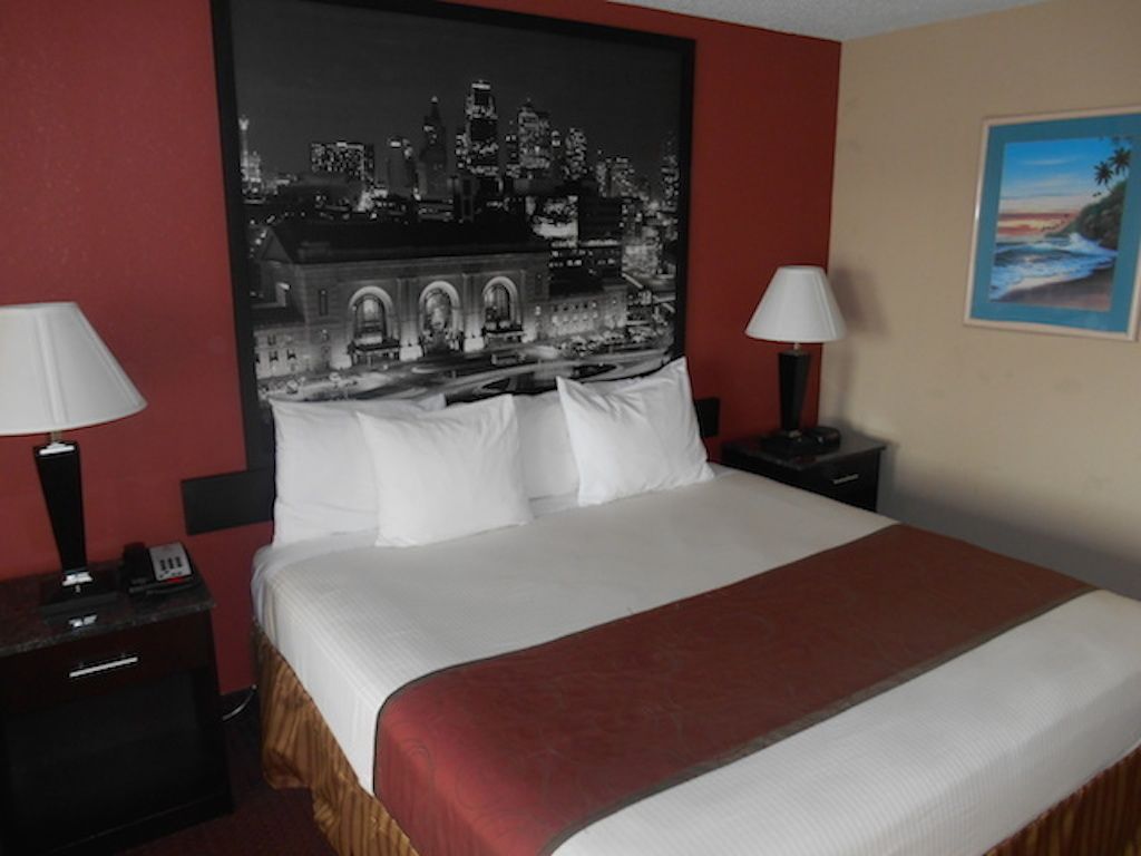 A hotel room with a large bed, two bedside tables, a cityscape artwork on the wall, and a painting of a beach scene.