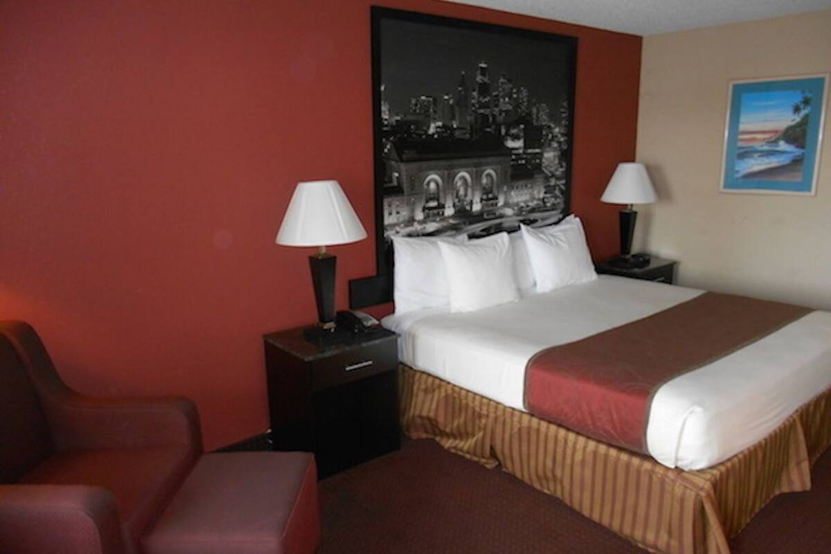 A hotel room with a large bed, two bedside tables with lamps, a red chair, and a cityscape painting on the wall.