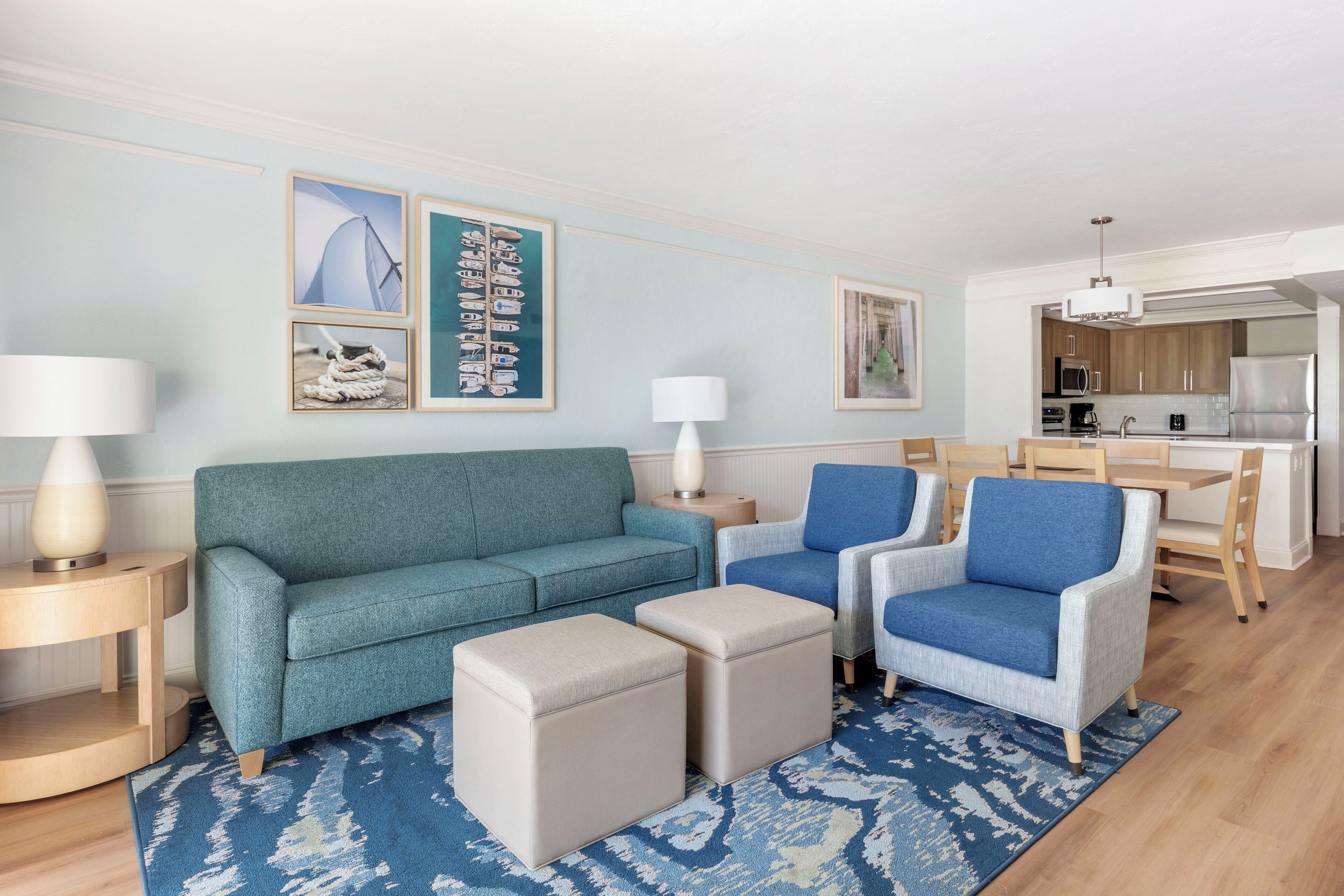A living room with a teal sofa, two blue armchairs, and a coffee table. There are framed pictures on the wall and a kitchen in the background.