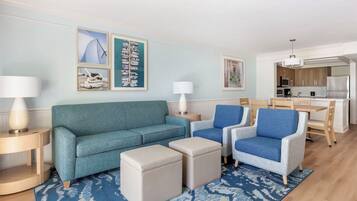 A living room with a teal sofa, two blue armchairs, and a coffee table. There are framed pictures on the wall and a kitchen in the background.