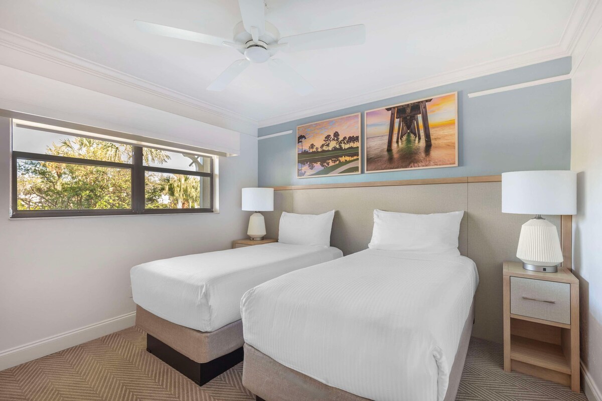 Une chambre d’hôtel avec deux lits, un ventilateur de plafond, une fenêtre donnant sur des arbres et des œuvres d’art accrochées au mur.