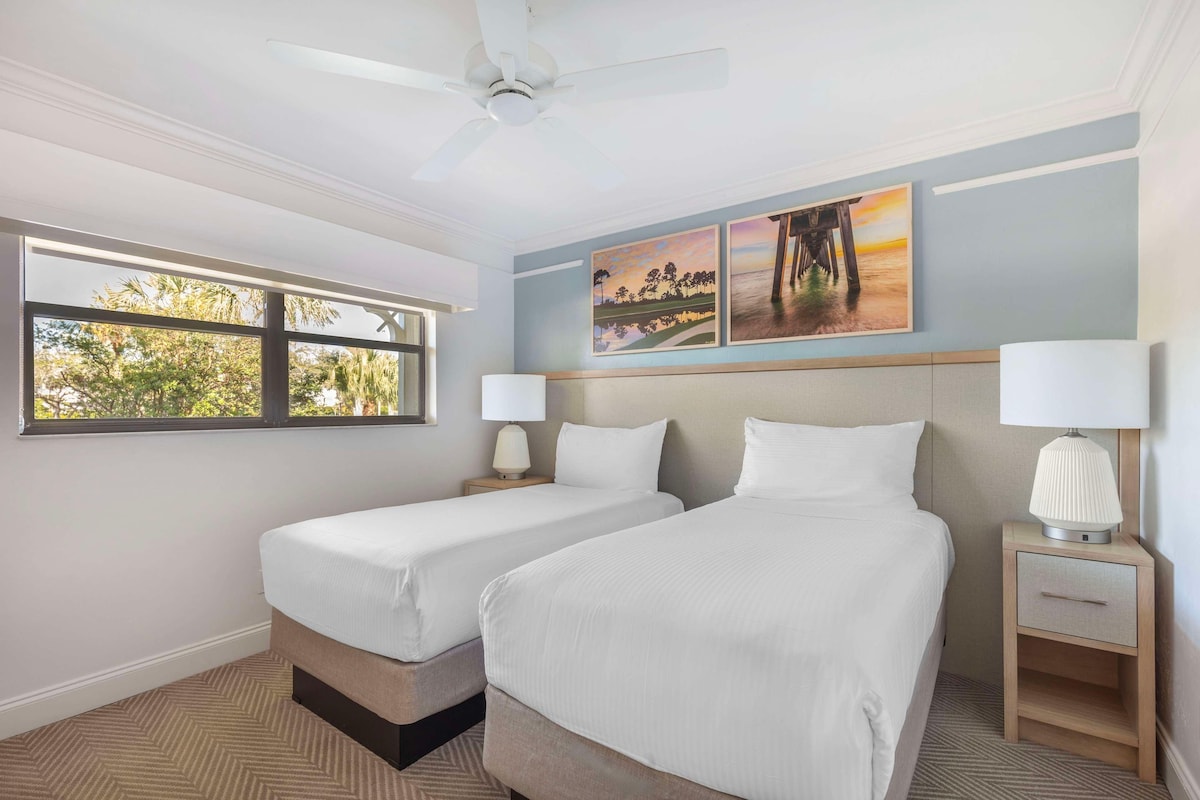 A hotel room with two beds, a ceiling fan, a window with a view of trees, and artwork on the wall.