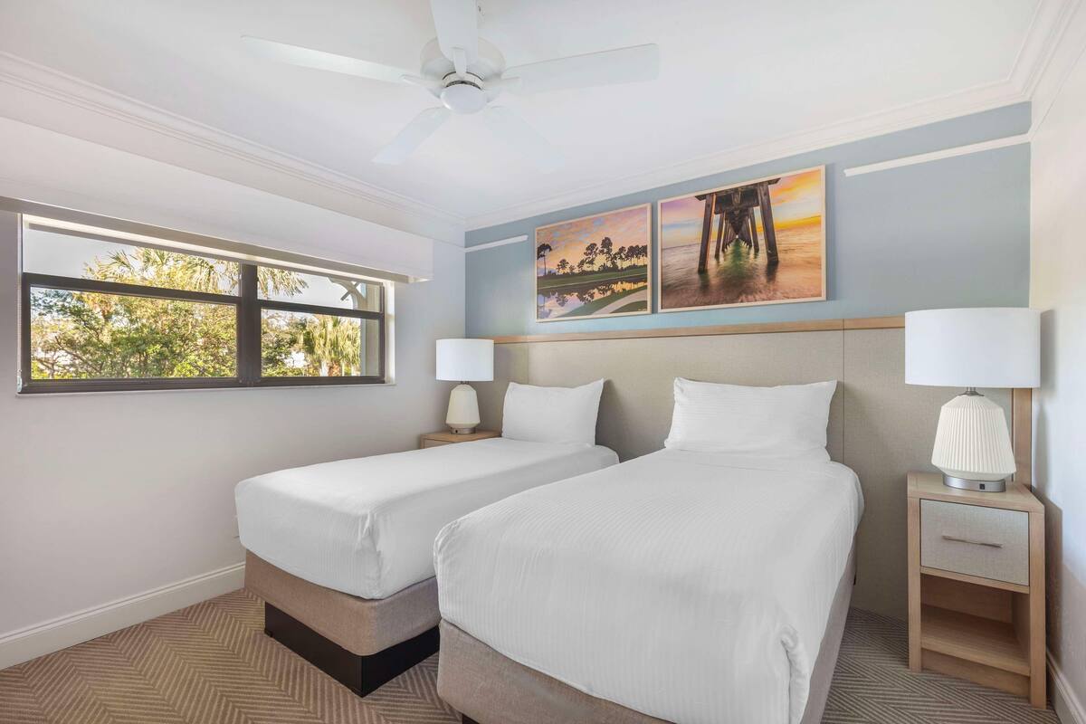 A hotel room with two beds, a ceiling fan, a window with a view of trees, and artwork on the wall.