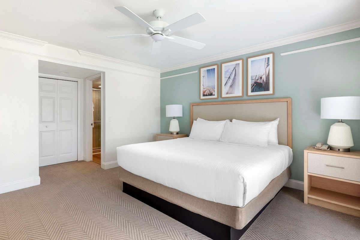 Une chambre d’hôtel avec un lit, des tables de chevet, un ventilateur de plafond et des tableaux encadrés au mur.