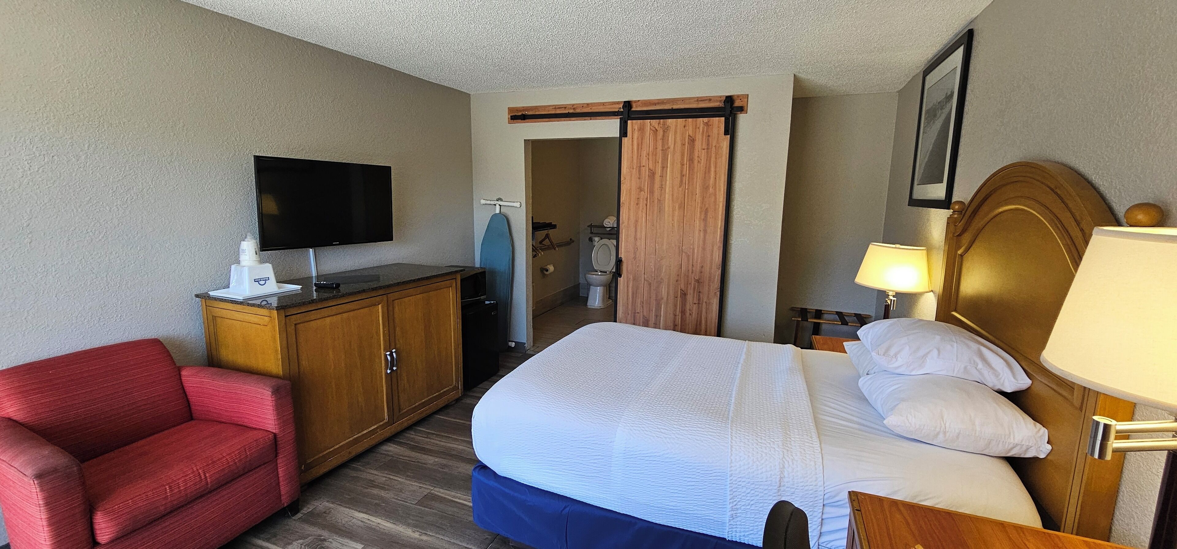 A hotel room with a bed, a TV, a desk, a chair, and a bathroom visible through a sliding door.