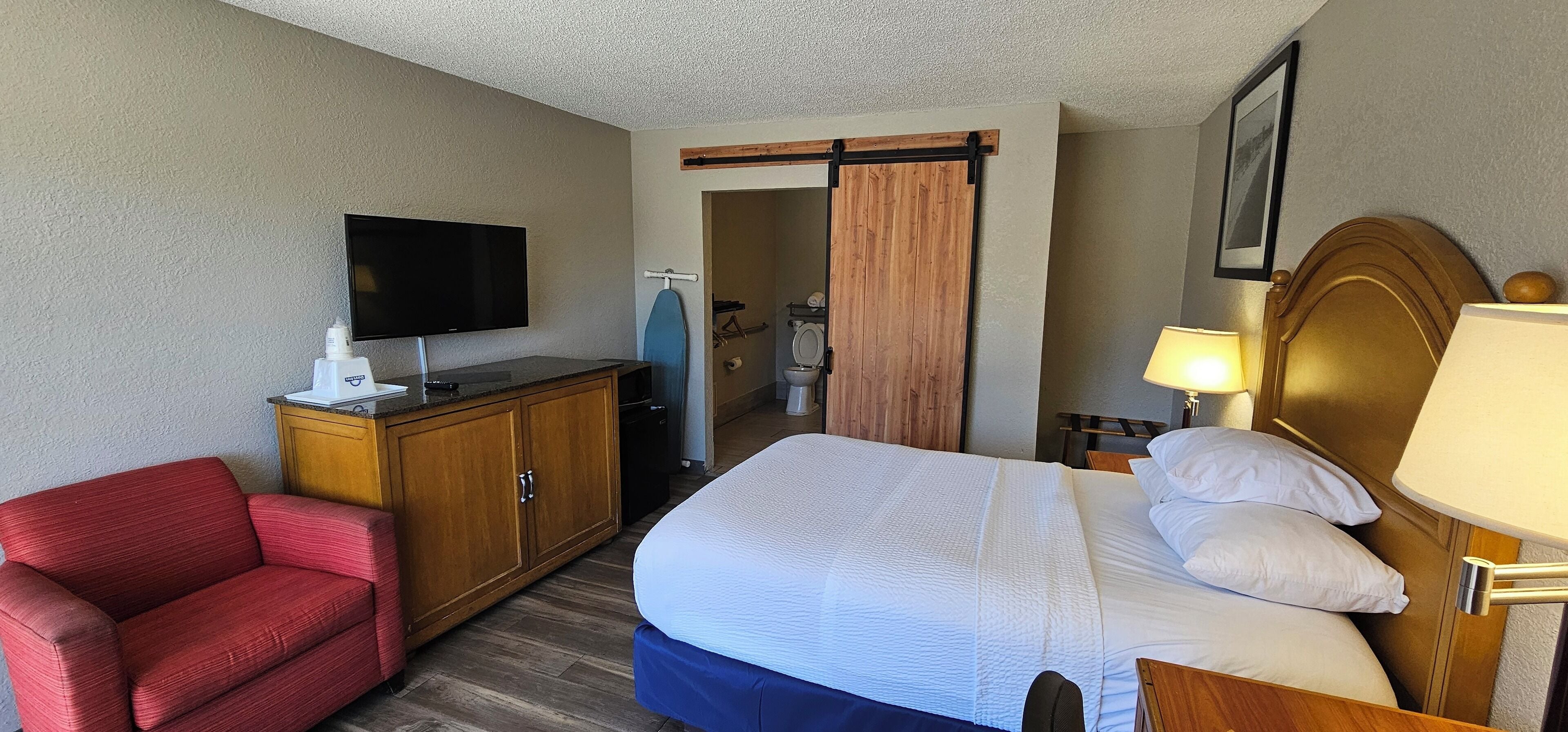 A hotel room with a bed, a TV, a desk, a chair, and a bathroom visible through a sliding door.