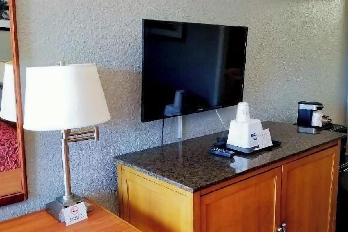 Quarto de hotel com uma cama, uma televisão na parede, uma cafeteira, um abajur e um pequeno armário de madeira.