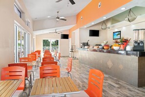 Ein Restaurant mit orangefarbenen Stühlen und Holztischen, einer Theke mit Speisen und Getränken sowie einem an der Wand befestigten Fernseher.