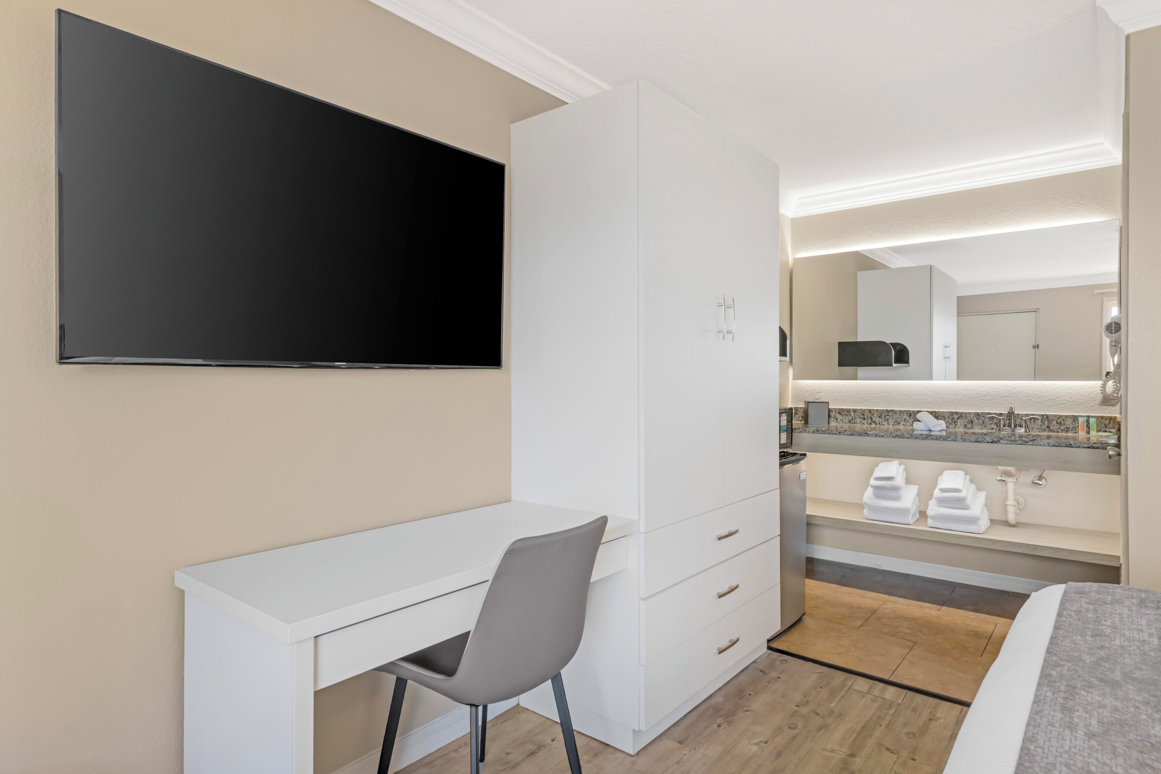 A hotel room with a flat-screen TV, a desk with a chair, a bed, a bathroom with a sink and mirror, and a minibar.