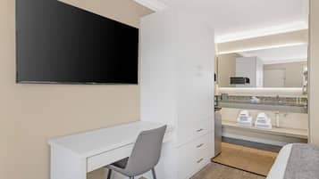 A hotel room with a flat-screen TV, a desk with a chair, a bed, a bathroom with a sink and mirror, and a minibar.