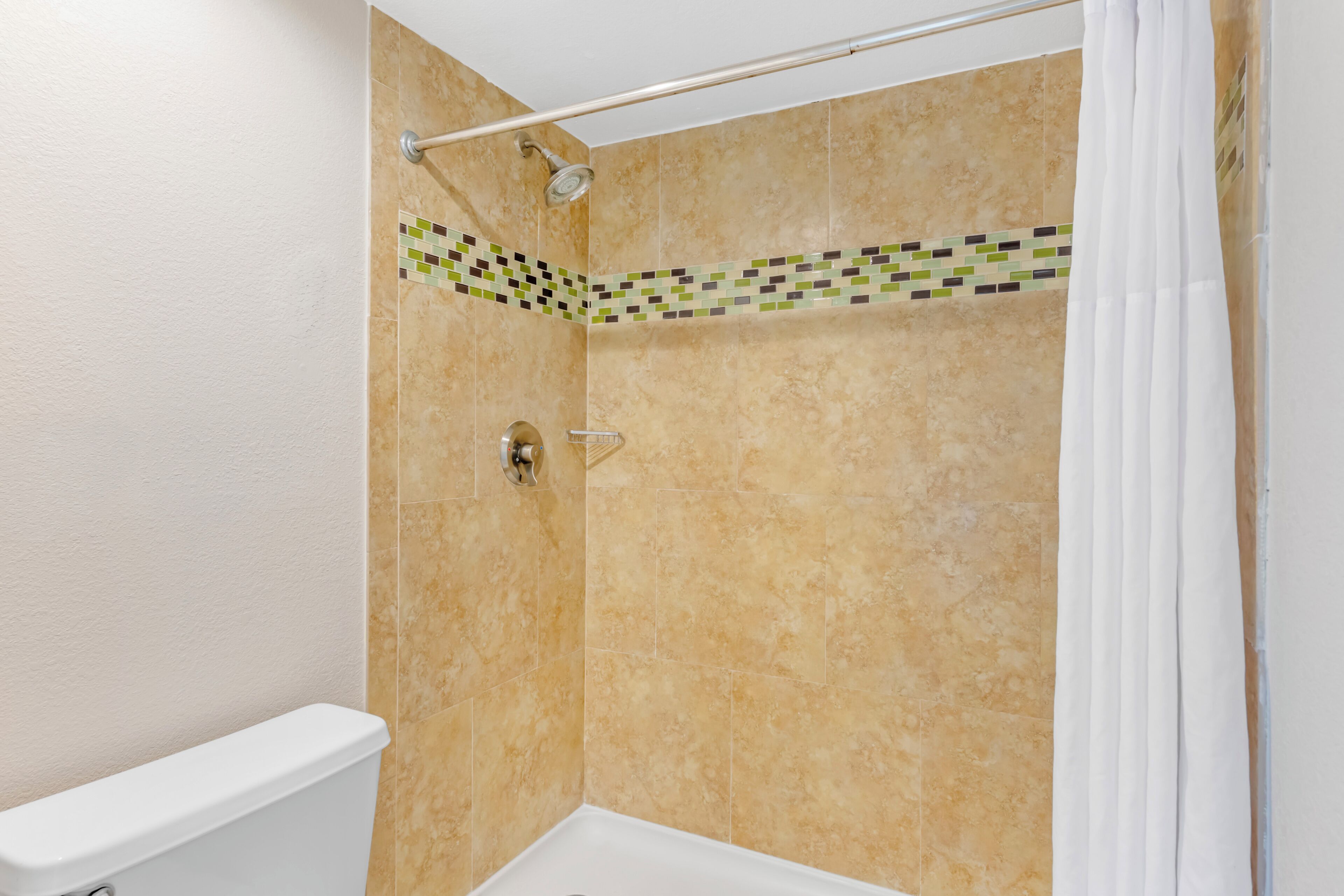 Une salle de bain avec des carreaux de couleur beige, une douche dotée d’un pommeau de douche à pluie, des toilettes blanches et un coin agrémenté d’une bordure en mosaïque.