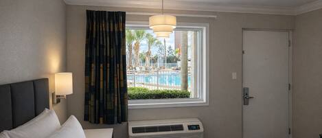 A hotel room with a bed, a window with curtains, a pool view, a door, and a wall-mounted air conditioner.