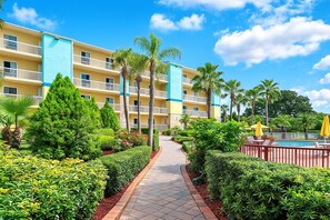 Un hotel con zona de piscina, un sendero bordeado de vegetación y un cielo azul despejado.