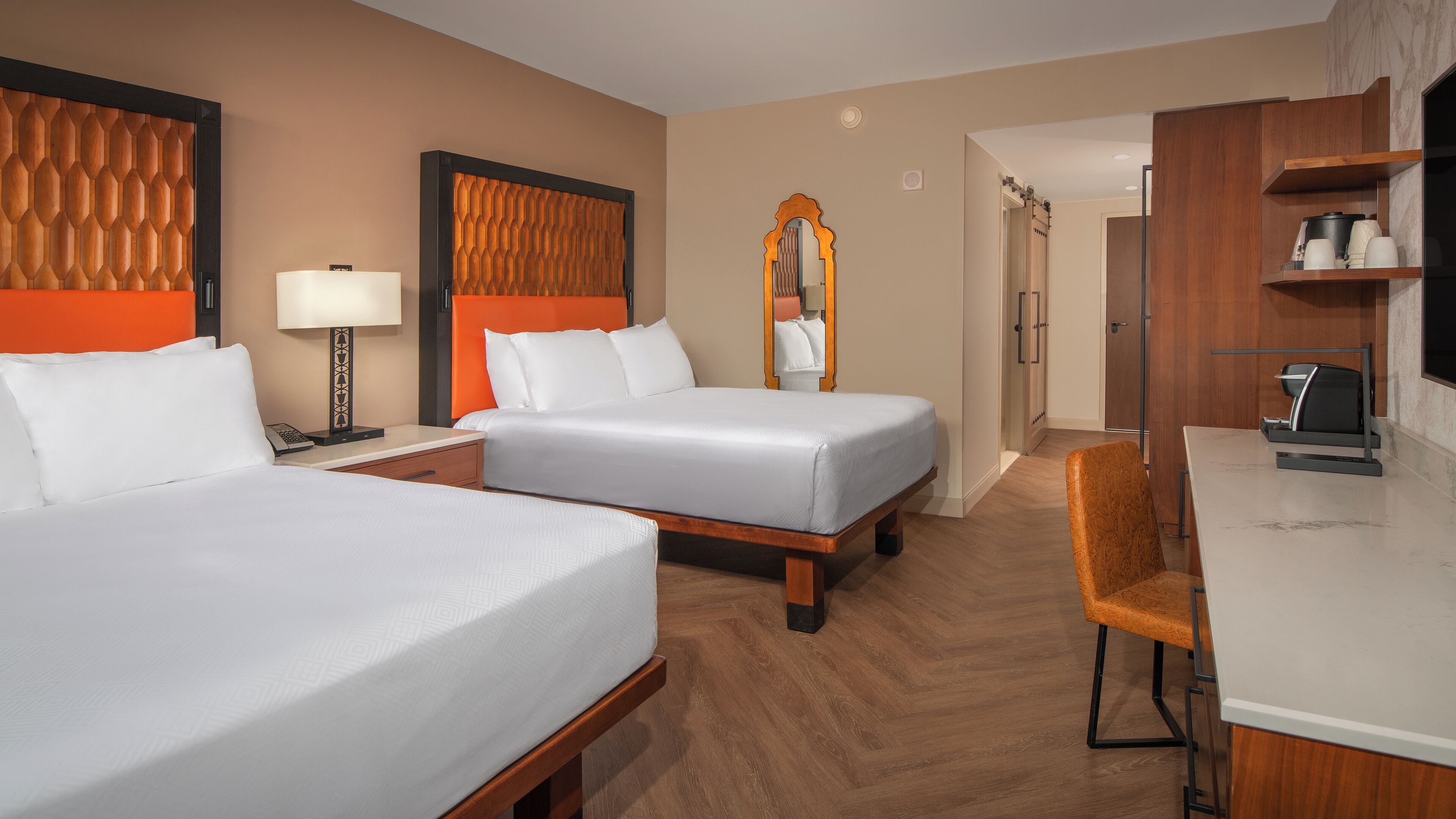 A hotel room with two beds, a desk, a chair, a mirror, and a coffee machine.