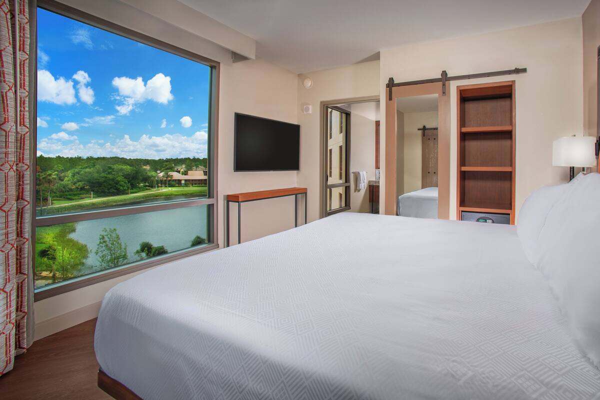 Een hotelkamer met een groot bed, een televisie, een houten kledingkast, een badkamer en uitzicht op een meer en bomen.