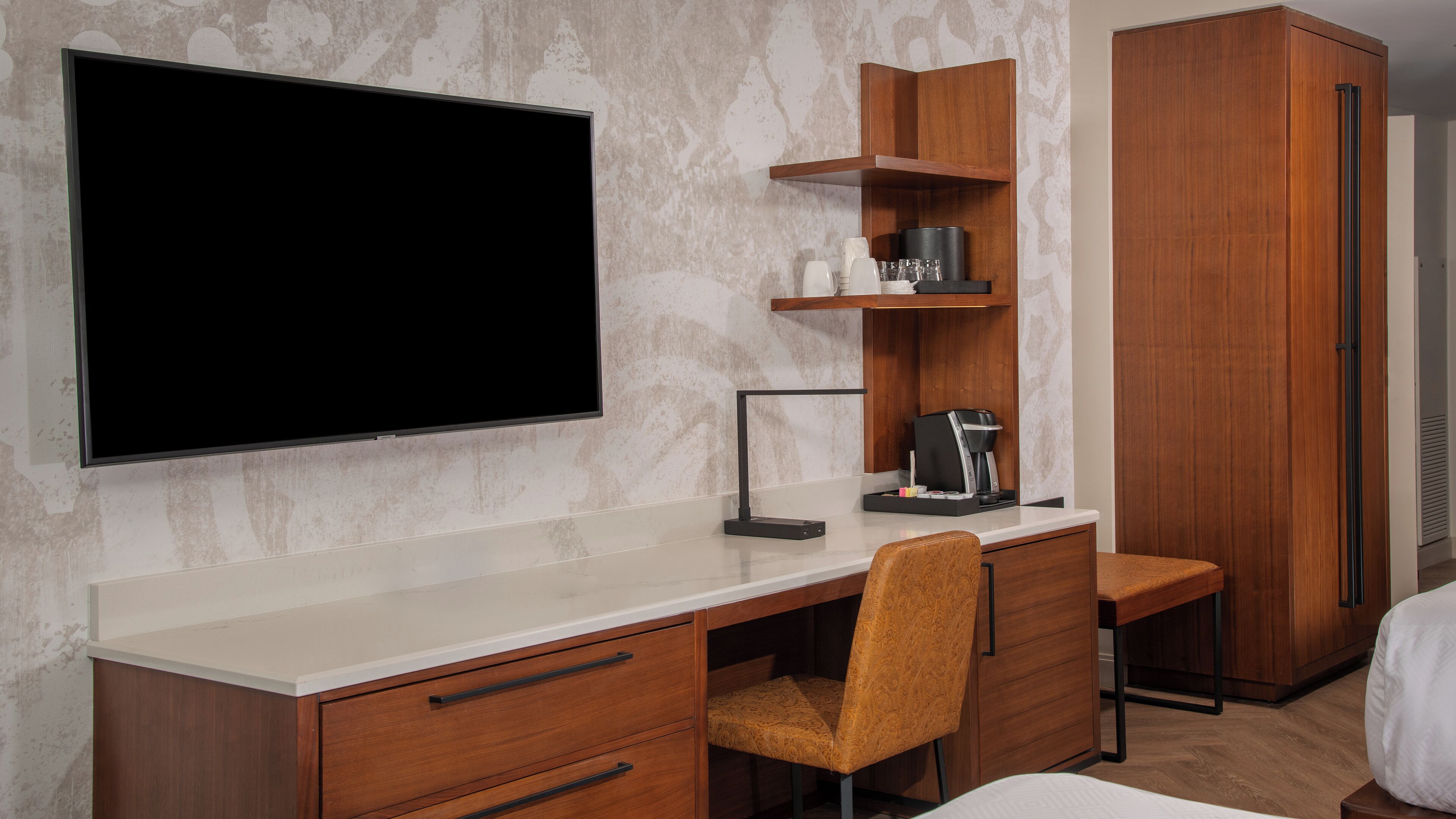 Une chambre d’hôtel moderne dotée d’un grand téléviseur à écran plat fixé au mur, d’un bureau en bois avec une machine à café, d’une chaise et d’une armoire.