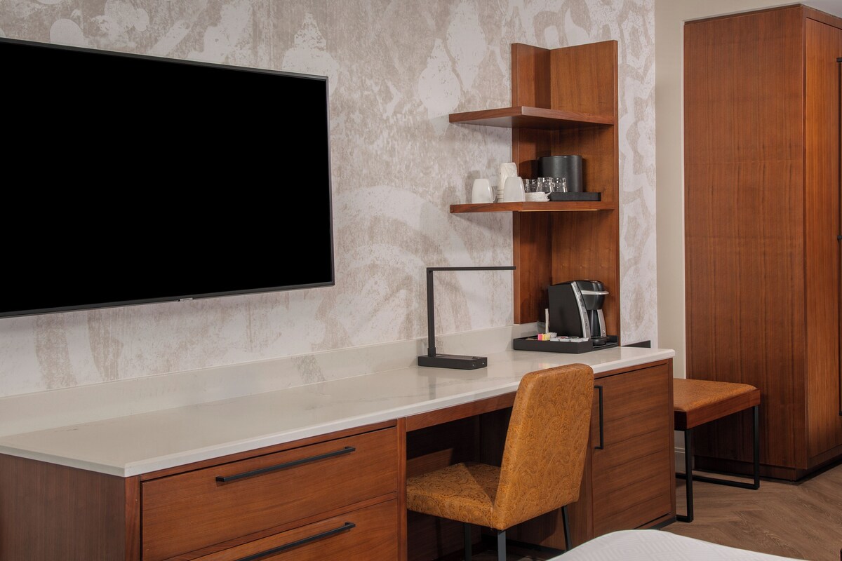 Una habitación de hotel moderna con un televisor de pantalla plana grande montado en la pared, un escritorio de madera con cafetera, una silla y un armario.