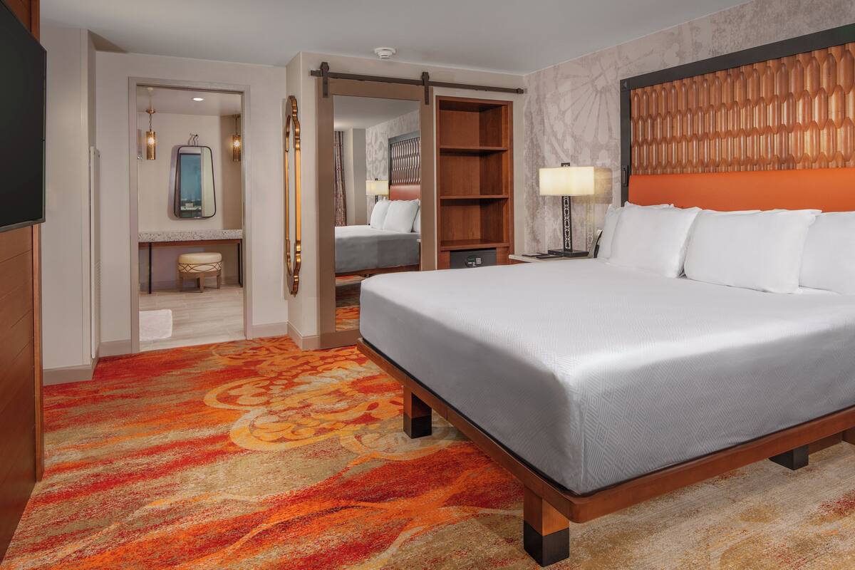 Hotelkamer met een groot bed, een houten hoofdbord, nachtkastje, bureau, stoel, garderobe, spiegel en een vloerbedekking.