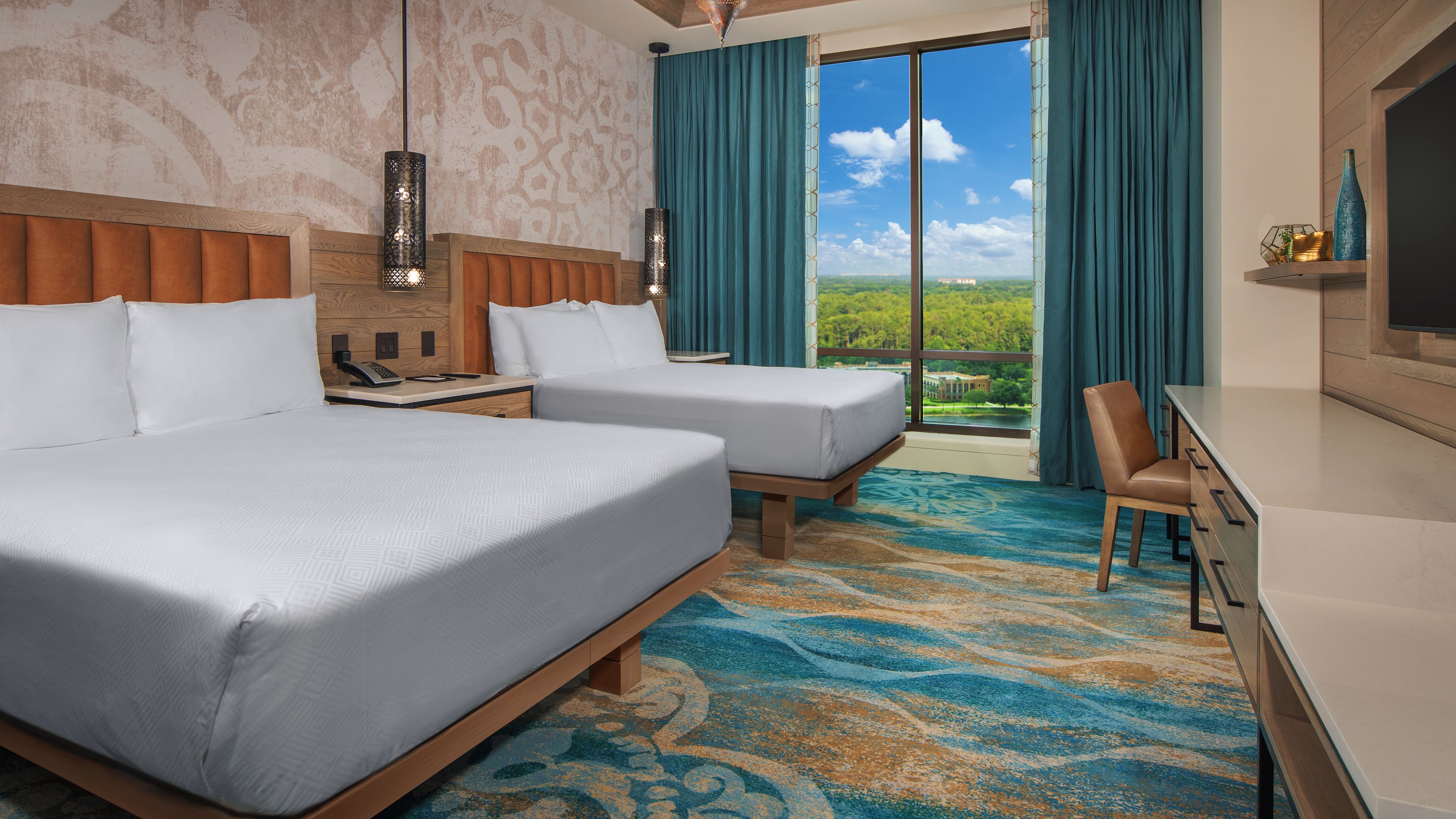 Hotelkamer met twee bedden, een bureau, een stoel, een televisie en een groot raam met uitzicht op groen.