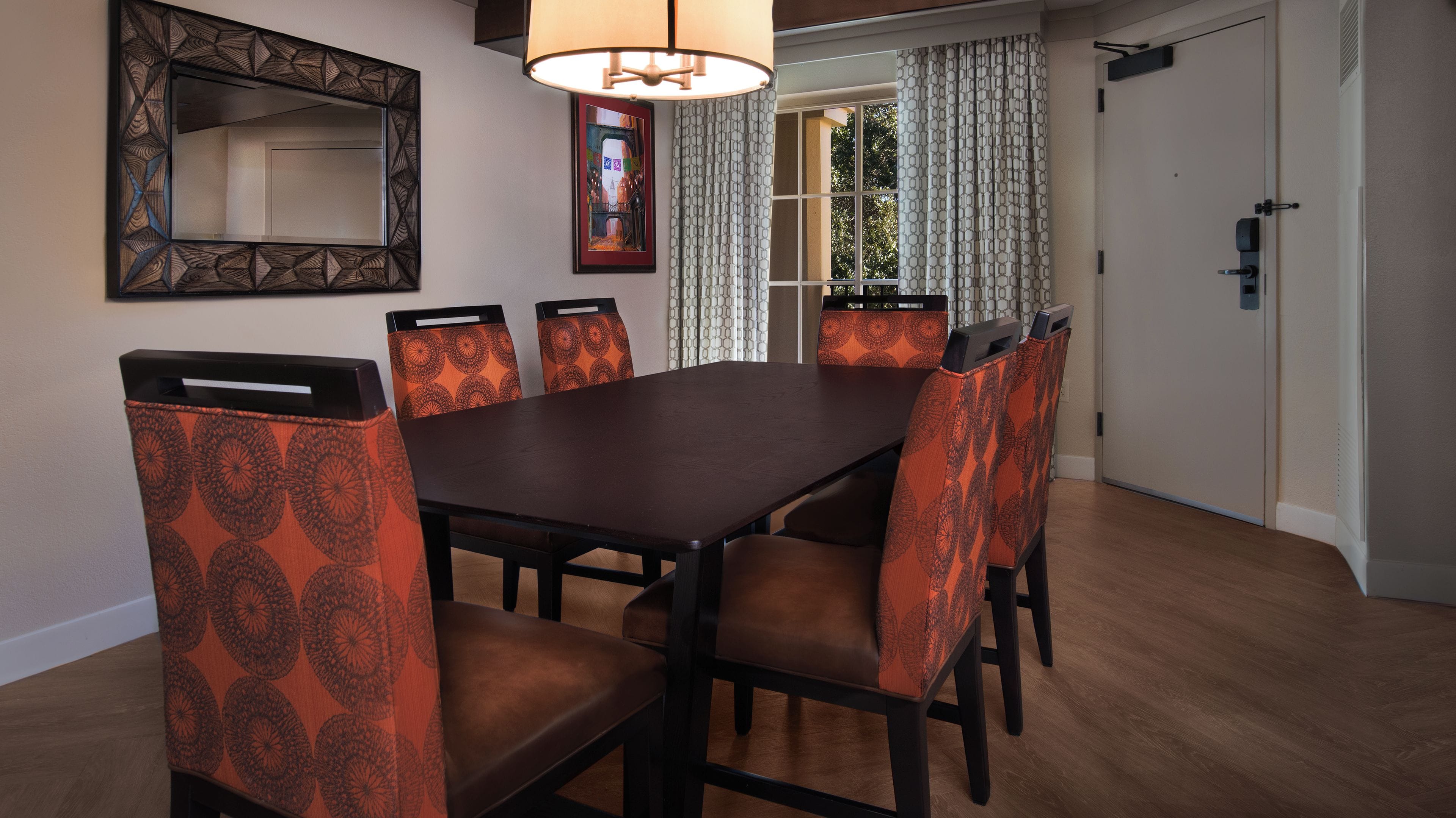 A dining area with a rectangular table surrounded by six chairs with patterned upholstery, a wall-mounted mirror, and a door with a patterned curtain.
