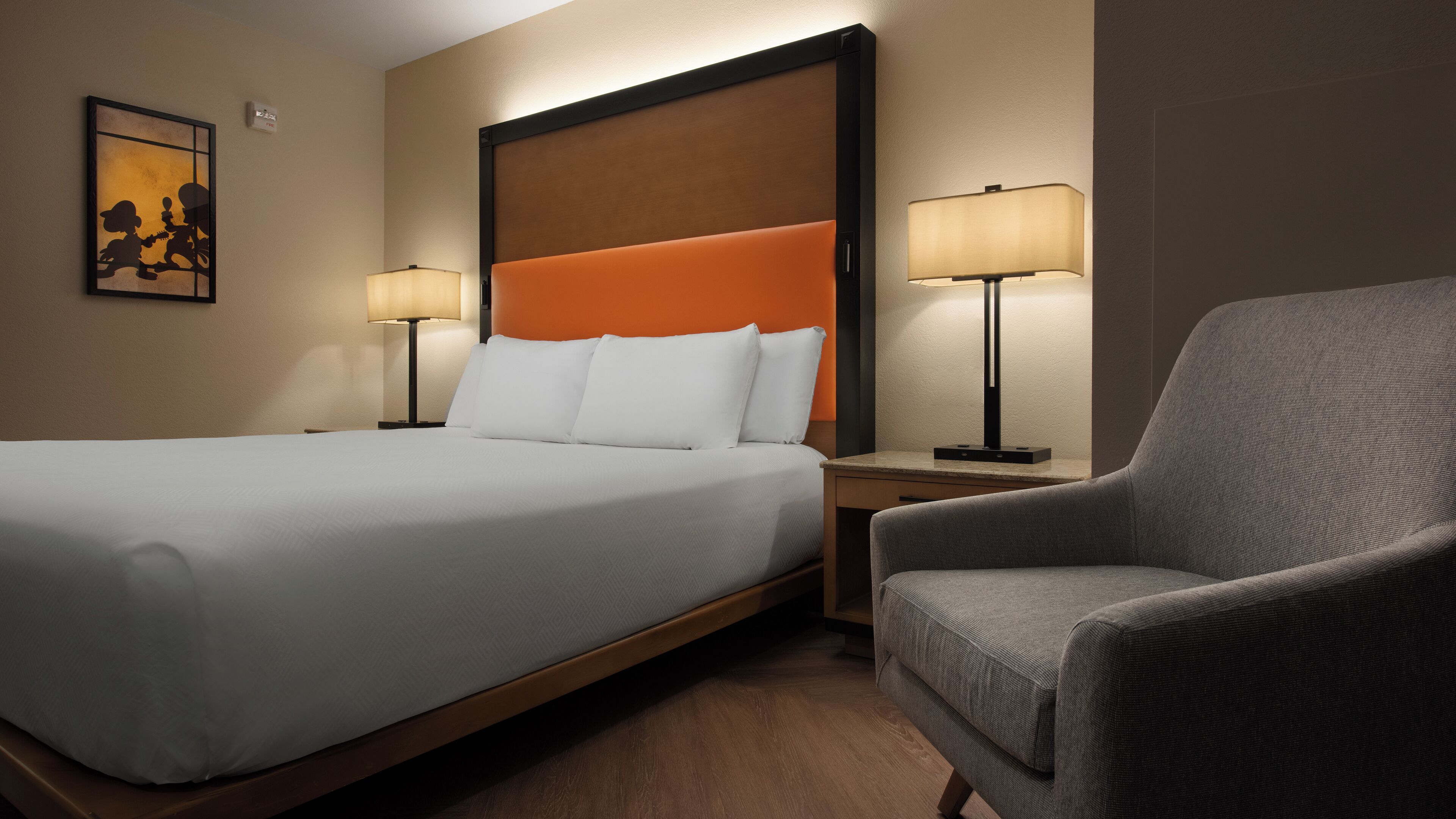 Une chambre d’hôtel avec un grand lit, deux tables de chevet avec des lampes, un fauteuil gris et un tableau encadré au mur.