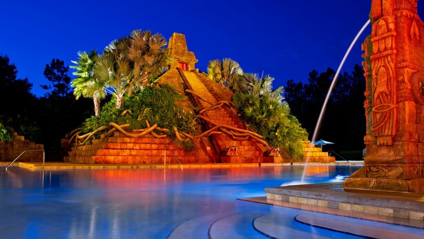 A nighttime view of a water feature with a stone structure resembling a temple, surrounded by palm trees and a pool. - Disney's Coronado Springs Resort (Lake Buena Vista)