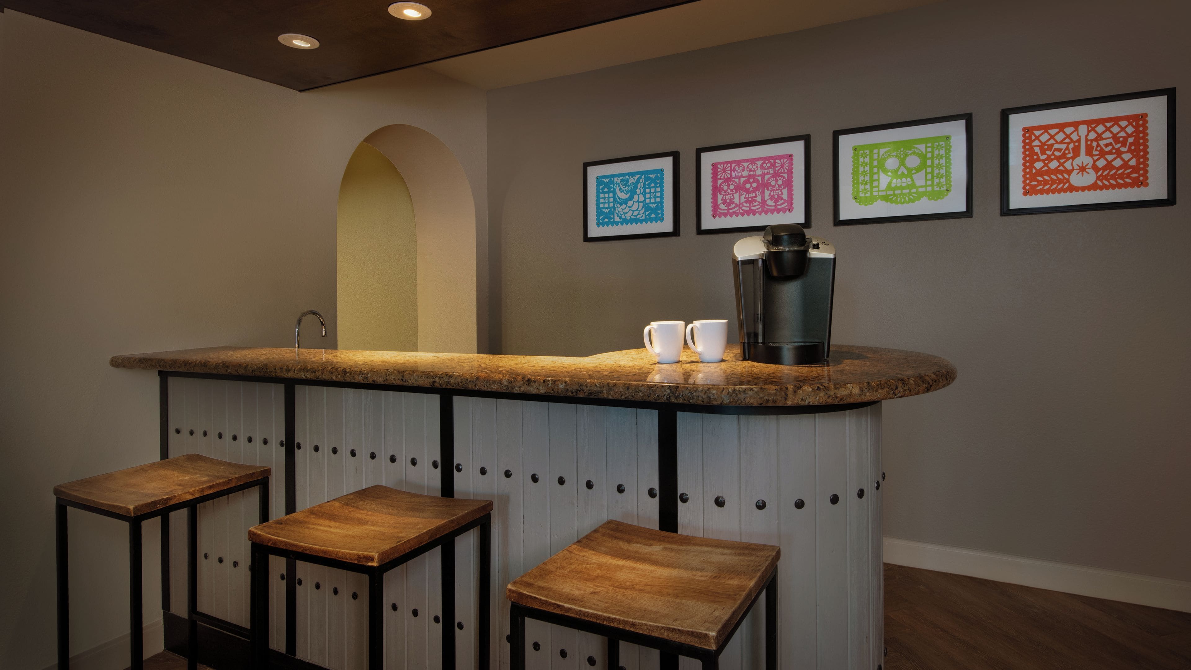 Una zona de bar con mostrador, dos taburetes, una máquina de café y cuatro obras de arte enmarcadas en la pared.
