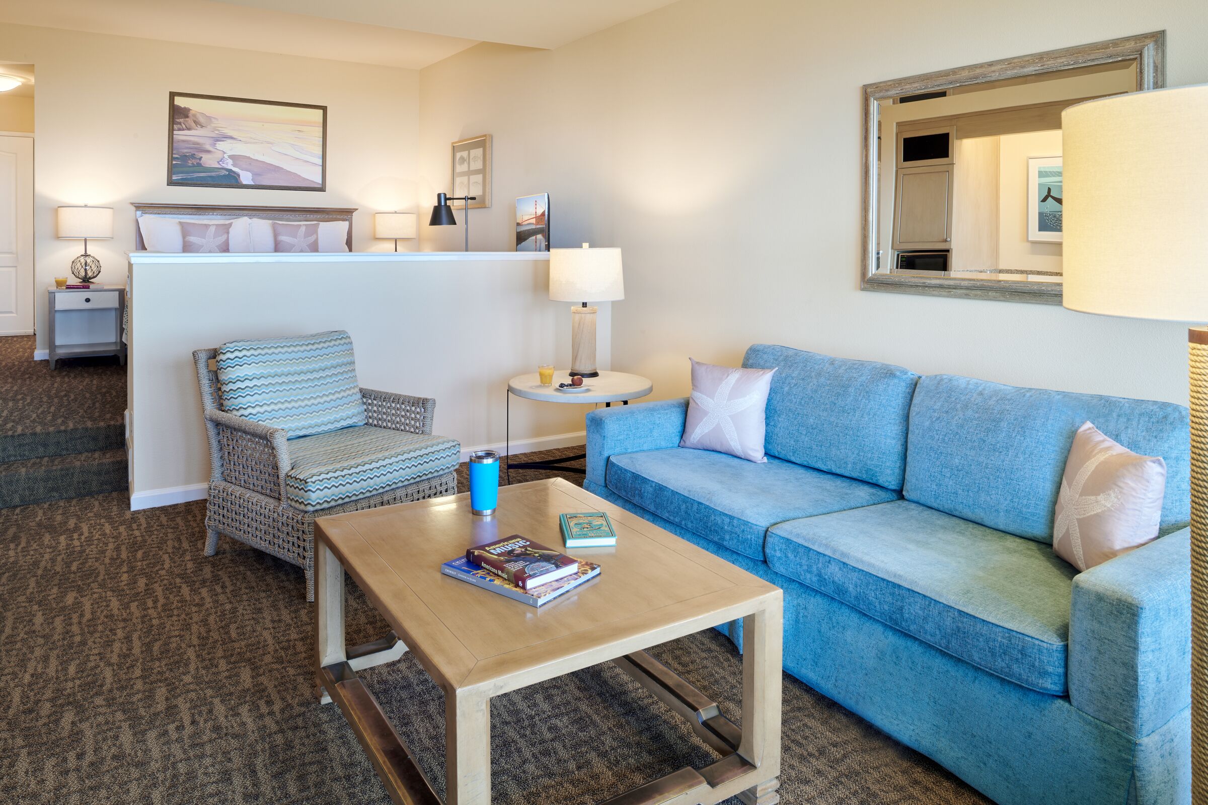 A hotel room with a blue sofa, a wicker chair, a wooden coffee table, and a small side table with a lamp.