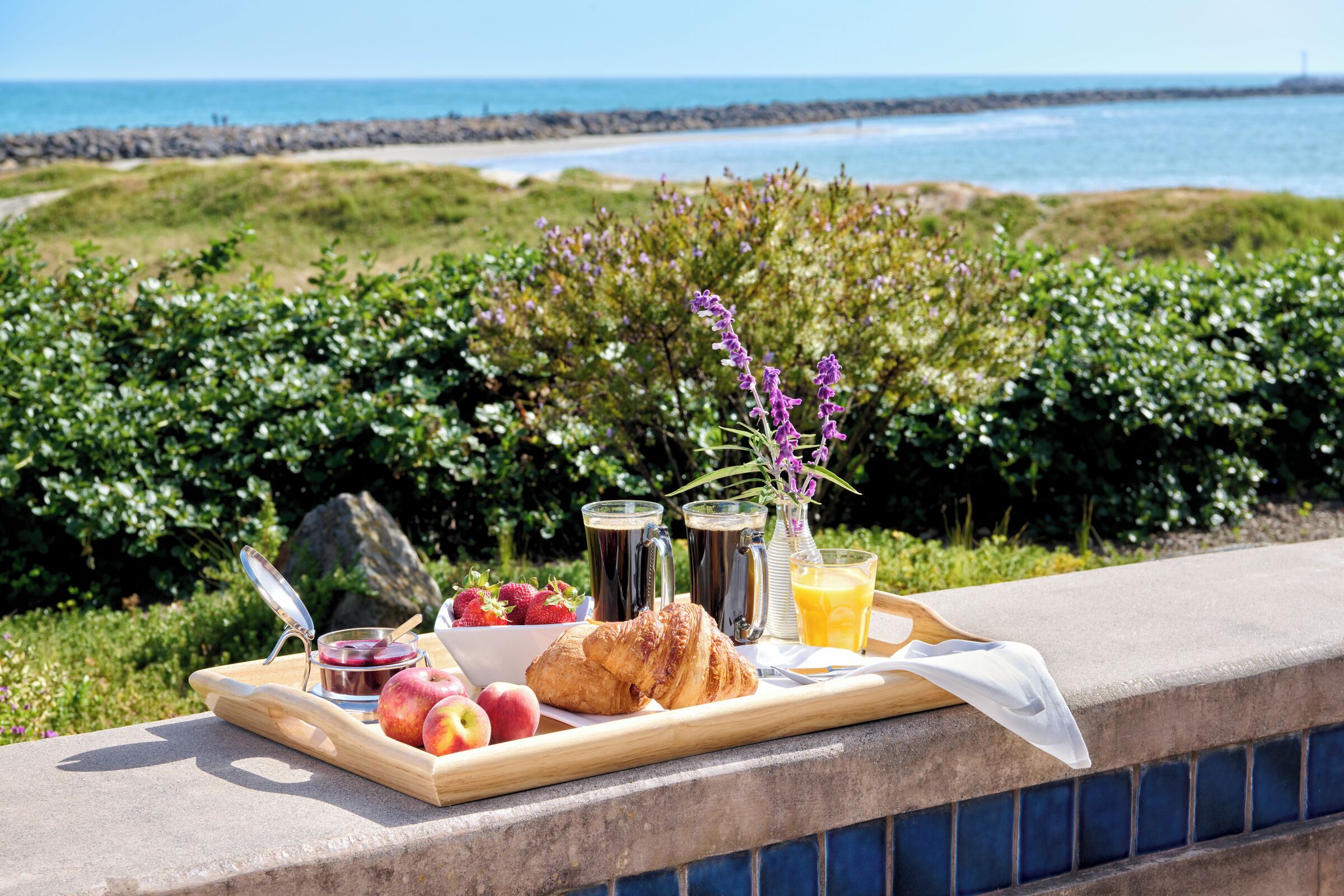 Un plateau de petit-déjeuner avec des croissants, des fraises, des pommes et un pot de confiture, posé sur une étagère donnant sur une plage.