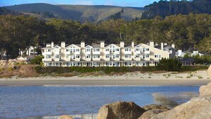 Een hotel aan het strand met meerdere verdiepingen, een zandstrand op de voorgrond en een bebost heuvel in de verte.