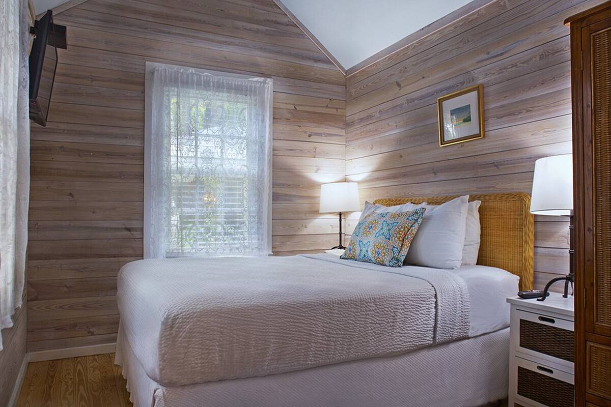 A bedroom with a wooden ceiling, a bed with white bedding, a nightstand with a lamp, and a window with sheer curtains.
