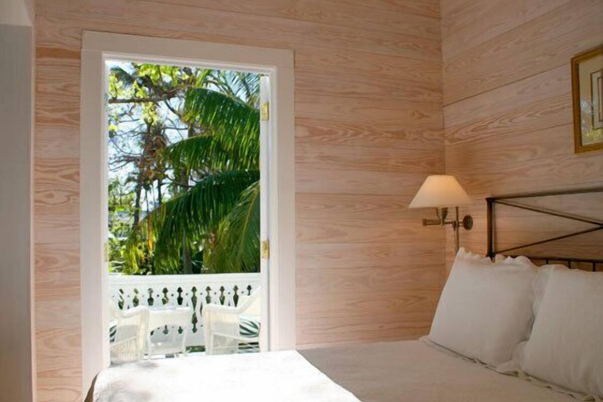 A bedroom with a bed, a nightstand, a lamp, and a view of palm trees through a door.